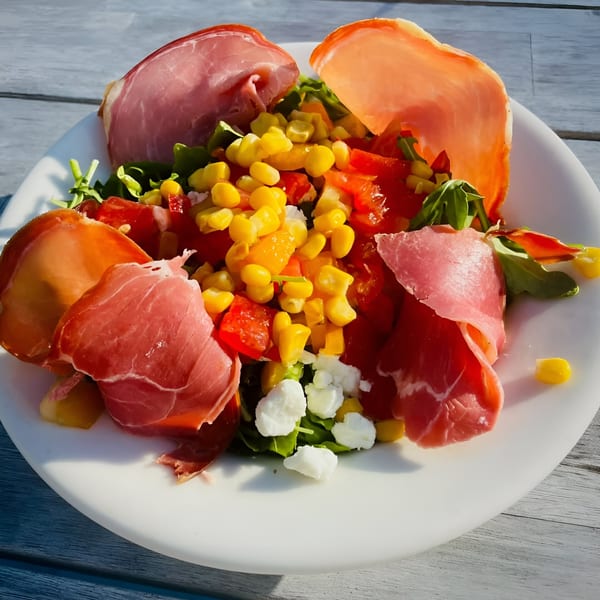 A fresh salad with prosciutto, corn, tomatoes, and feta cheese on a white plate.