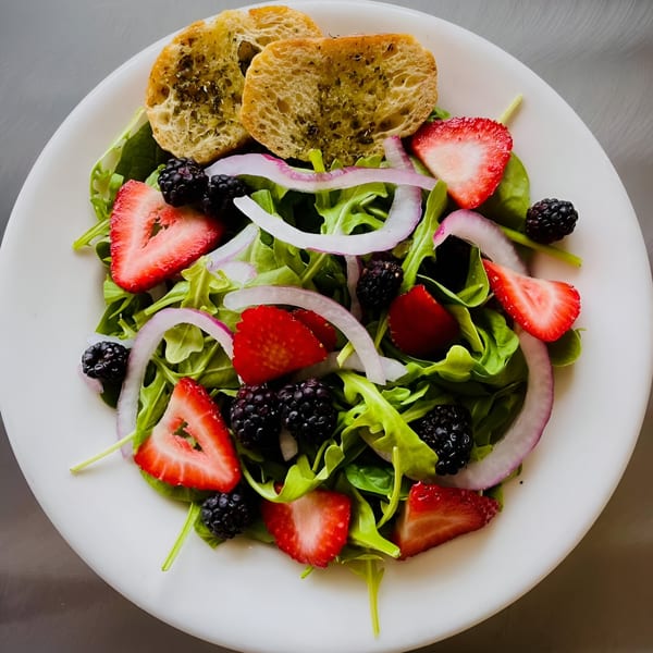 A fresh salad with arugula, strawberries, blackberries, red onion, and garlic bread on a white plate.
